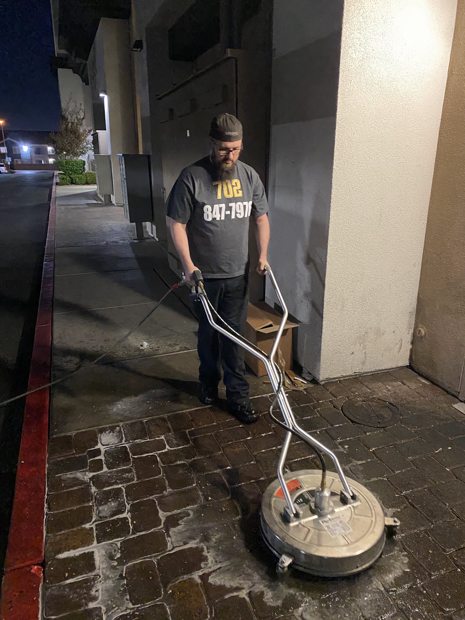 Worker in a branded 702 shirt using a surface cleaner on a paver walkway at night